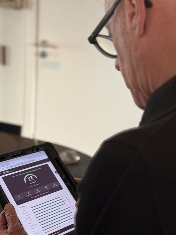 Een bewoner bekijkt over zijn schouder de VeriCare-app op een tablet: een woningscore van 93 met de lijst van controlepunten eronder.