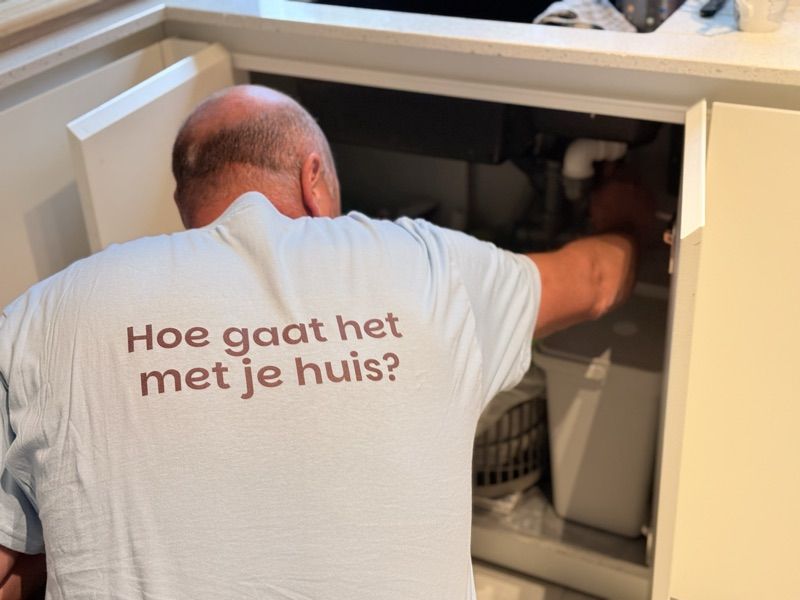 Een VeriCare-medewerker, shirt met de tekst "Hoe gaat het met je huis?", herstelt de afvoer in het keukenkastje onder de gootsteen.