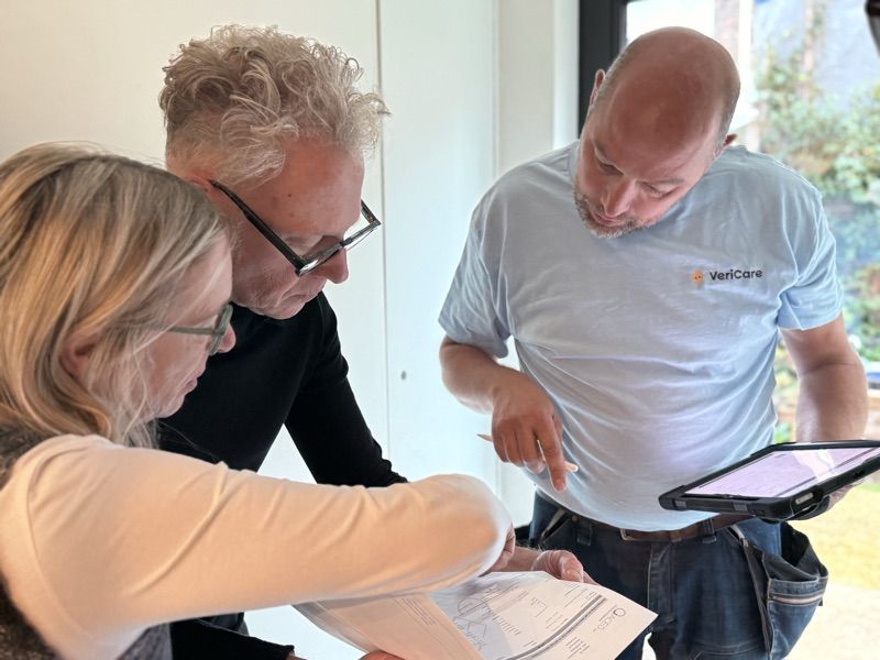Een VeriCare-inspecteur in lichtblauw shirt toont een koppel de controlelijst op een tablet en op papier, binnen in hun woning.