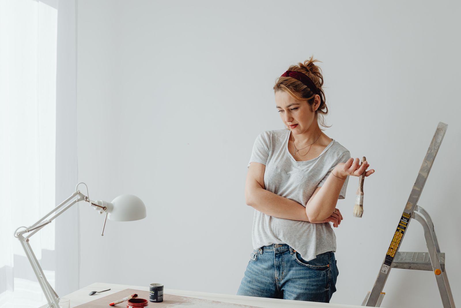 Vrouw met kwast in de hand kijkt twijfelend naar de muur, ladder en gereedschap ernaast.